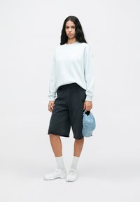 Γυναίκα με μακριά μαύρα μαλλιά φοράει ανοιχτό μπλε φούτερ, μαύρα σορτς μέχρι το γόνατο, λευκά παπούτσια και κάλτσες, κρατώντας ένα ανοιχτό μπλε μπέιζμπολ καπέλο.