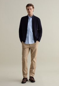 Maglione zip-up blu navy sopra una camicia a righe blu chiaro e bianca, pantaloni chinos beige e scarpe marroni, esposto su uno sfondo neutro.