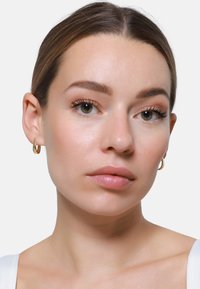 Orecchini a cerchio in metallo dorato, con una superficie liscia e lucida. Il volto della modella presenta un trucco naturale e un tono di pelle neutro.