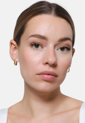 Oorbellen in de vorm van hoops, gemaakt van goudkleurig metaal, met een gladde, gepolijste oppervlakte. Het gezicht van het model is voorzien van natuurlijke make-up en een neutrale huidtint.