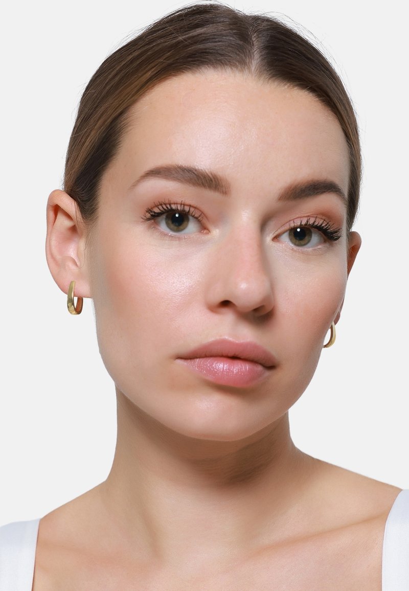 Orecchini a cerchio in metallo dorato, con una superficie liscia e lucida. Il volto della modella presenta un trucco naturale e un tono di pelle neutro.