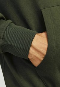 Sötétzöld pulóver anyag bordázott mandzsettákkal, tenyér a zsebben pihen, textúrált felülettel és letisztult varrási részletekkel.
