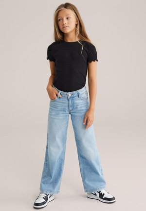 Mladé dievča s dlhými vlasmi, oblečené v čiernom priliehavom tričku, svetlomodrých širokých džínsoch a čierno-bielych teniskách, stojace s jednou rukou v vrecku.