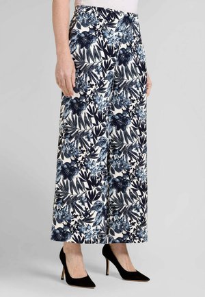 Donna che indossa pantaloni a gamba larga con stampa floreale nera, bianca e blu e scarpe nere con tacco alto, in piedi su uno sfondo grigio.