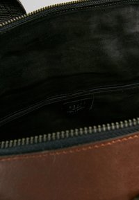 Hnědá kožená taška uvnitř s měkkým černým podšívkou, obsahující kapsu na zip a štítek. Pevný zip a úhledné detaily šití.