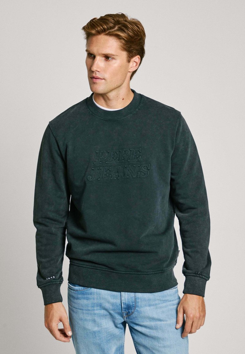 Sudadera verde oscura con el logo grabado de "PEPE JEANS", cuello y puños acanalados, y una textura suave, combinada con jeans de color azul claro.