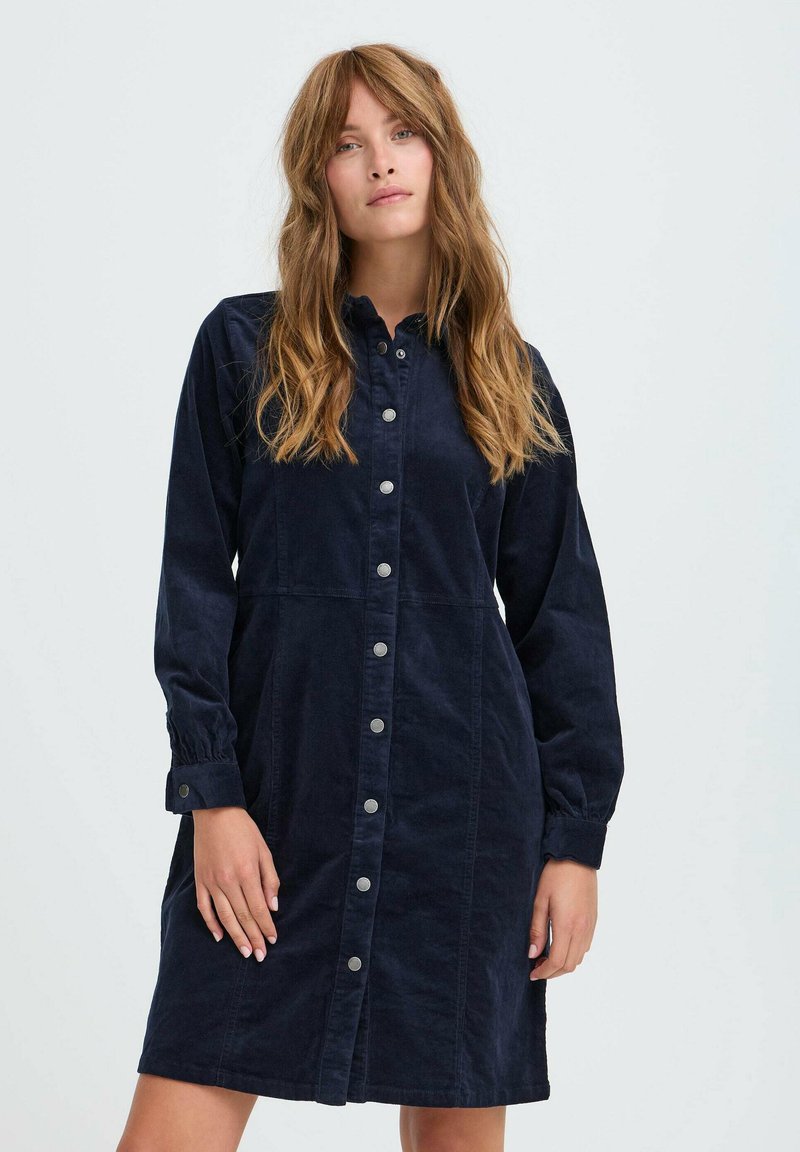 Kvinde med langt bølget hår iført en marineblå knappet langærmet kjole, stående mod en ensfarvet lys baggrund.