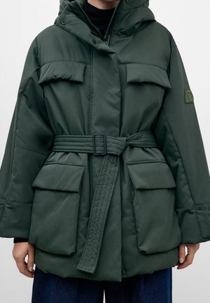 Dunkelgrüne gesteppte Jacke mit Gürtel, großen Klappentaschen und Kapuze, getragen über schwarzem Oberteil und blauer Hose.