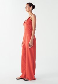 Žena stojaca bokom, oblečená v oranžovom overal s širokými nohavicami a tenkými ramienkami, v čiernych sandáloch, na pozadí bielej steny.