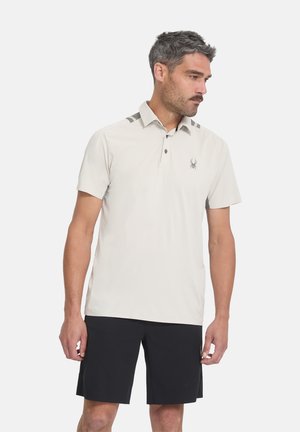 Homme portant un polo beige clair à manches courtes avec un logo araignée et un short sombre jusqu’aux genoux, debout devant un fond blanc uni.
