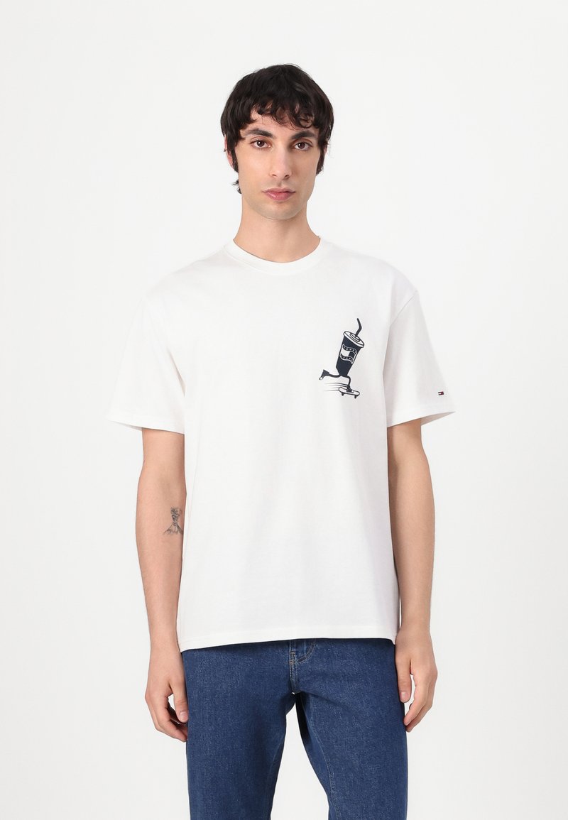 Una camiseta de algodón blanca presenta un gráfico de una bebida con una pajita en el lado izquierdo del pecho. El diseño es simple, con mangas cortas y un cuello redondo.