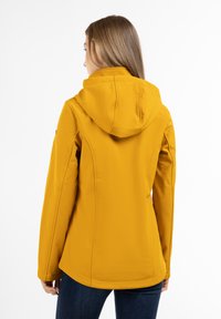 Gul softshell-jakke med hætte, der har sidesømmer og en glat tekstur. Båret over mørke jeans, der viser en tætsiddende silhuet.
