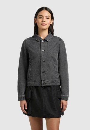 Femeie purtând o jachetă de denim neagră, decolorată, cu nasturi, peste o fustă neagră cu buzunare cargo, stând în fața unui fundal simplu, gri deschis.