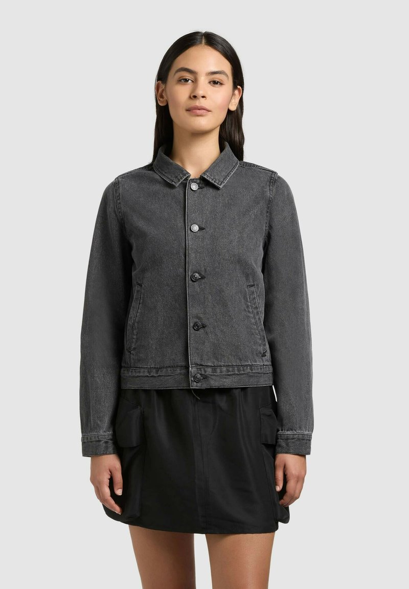 Femme portant une veste en jean noire délavée boutonnée par-dessus une jupe noire avec des poches cargo, debout devant un fond gris clair uni.
