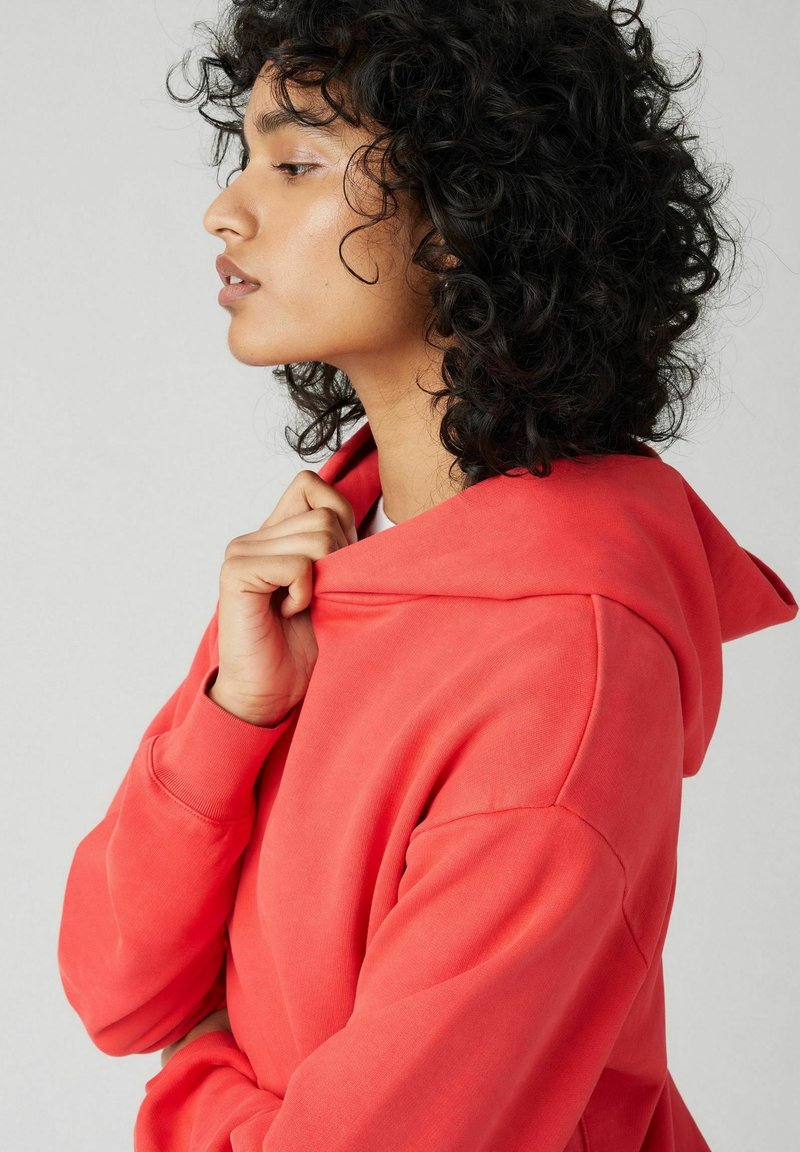 Korallfarbene Kapuzenpullover aus weichem Material mit langen Ärmeln, einem lockeren Schnitt und einer Kordelzugkapuze.