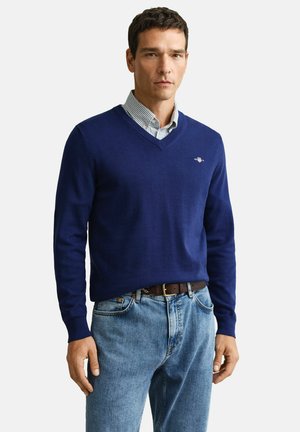 Man wearing a blue V-neck sweater over a striped button-up shirt, light blue jeans, and a brown belt, standing against a white background.