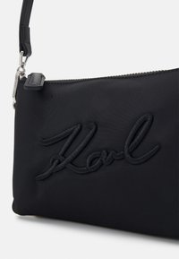 Sac d’épaule en tissu noir avec le texte "Karl" brodé, fermeture éclair et bandoulière amovible avec quincaillerie argentée.
