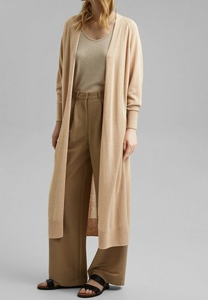 Femme portant un long cardigan beige sur un haut beige et un pantalon large marron avec des sandales noires ouvertes aux orteils, debout sur un fond gris uni.