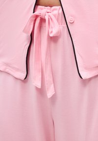 Κοντινή λήψη ροζ παντελονιού με δεμένη ζώνη υφάσματος στη μέση και ταυτόχρονα ροζ μπλούζα με μαύρο περιθώριο και λεπτομέρεια με κουμπί.