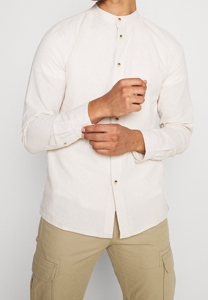 Homme portant une chemise blanche à col mandarin à manches longues en train de boutonner le poignet, avec un pantalon cargo beige, sur un fond uni.