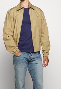 Muž v béžové mikině na zip s námořnickou košilí a světle modrými džíny, stojící s jednou rukou v kapse na jednobarevném pozadí.