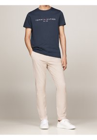 Granatowa bawełniana koszulka z białym nadrukowanym logo, zestawiona z jasnobeżowymi spodniami slim-fit i białymi sneakersami. Minimalistyczny design i wygodne dopasowanie.