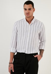 Camicia bianca a righe con linee navy, vestibilità rilassata, maniche lunghe arrotolate ai polsini. Abbinata a pantaloni neri, che mostrano un look informale.
