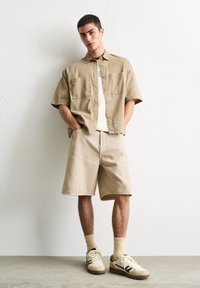 Beige Kurzarmhemd mit Knöpfen, passende Shorts und beige Sneaker mit braunen Akzenten. Getragen mit hellen Socken.