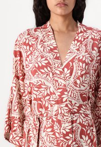 Raudonas raštuotas kimono su baltais gėlių motyvais, turintis gilų V formos iškirpimą, plačias rankoves ir nėrimo diržą ties juosmeniu. Audinys atrodo tekstūruotas.