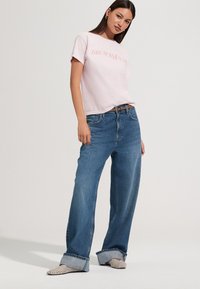 Svijetloružičasta majica s natpisom „BRUNCH&YARD“, uparena s širokim plavim trapericama s presavijenim manšetama i svjetlim uzorkovanim cipelama.