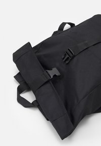 Mochila negra con material de tela, correas ajustables y cierre de hebilla de plástico. El diseño es estructurado con un estilo de rollo en la parte superior.