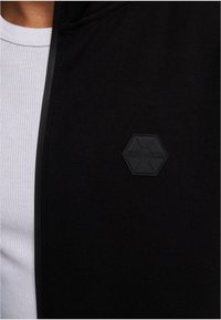 Sort zip-up hoodie med en struktureret sort hexagonal lap med teksten "SOUTHPOLE" på brystet. Underneath ses en hvid ribbet skjorte.