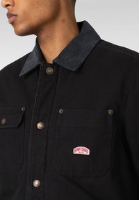 Svart bomullsjakke med mørk kordfløyelskrage, to frontlommer med messingsknapper og en rød etikett på venstre lomme.