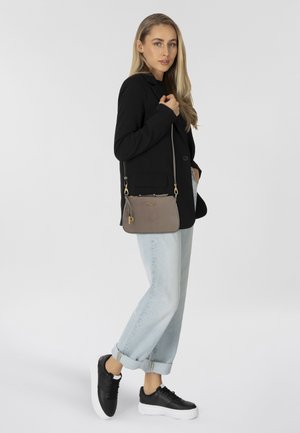 Graue Umhängetasche mit goldenen Akzenten, glatter Textur und Logodetail. Das Model trägt ein schwarzes Blazer und hochgerollte helle Jeans mit schwarzen Sneakern.