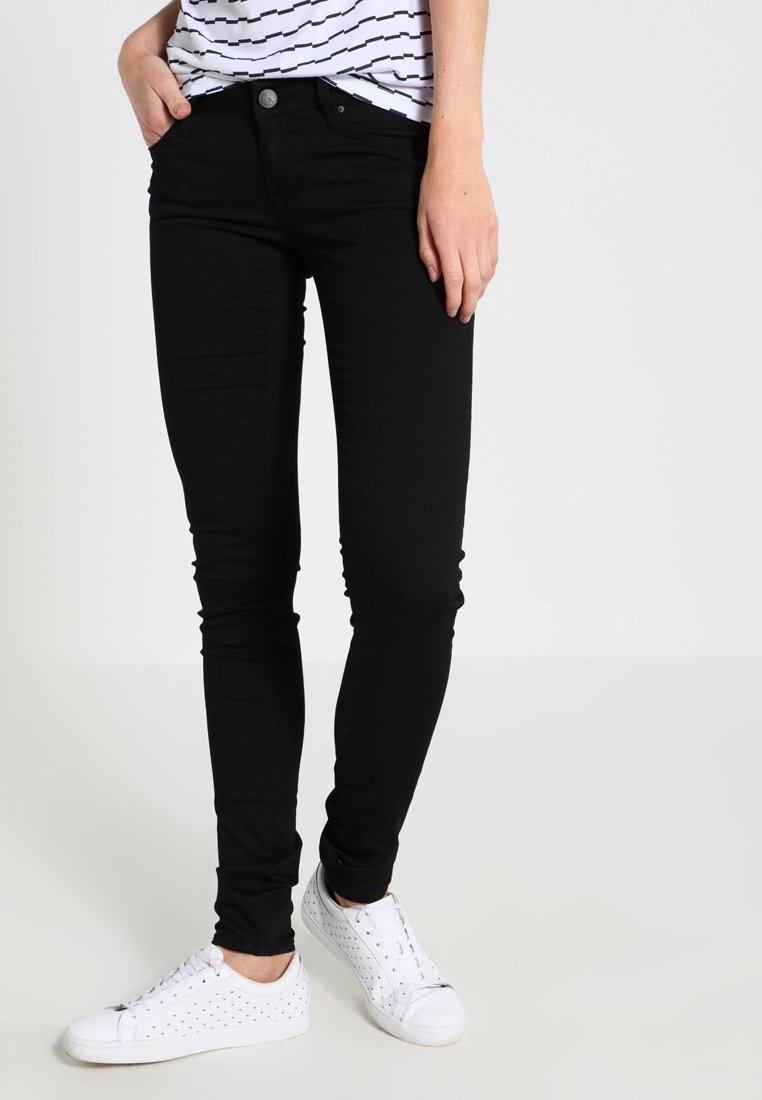 Pantalones negros de corte ajustado combinados con zapatillas blancas perforadas y una camisa de rayas de manga corta, persona de pie con una mano en el bolsillo.