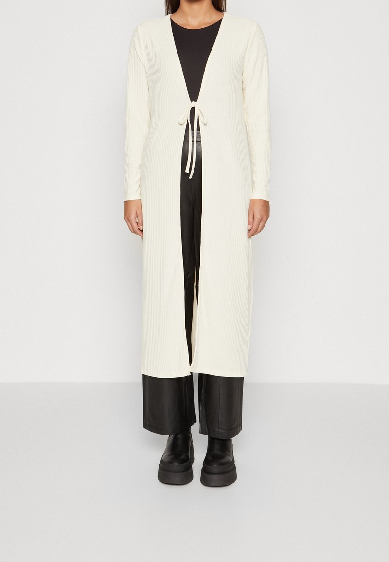 Femme portant un long cardigan côtelé crème noué à la taille, un top noir, un pantalon large en cuir noir et des bottes à plateforme noires.