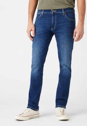 Jeans de mezclilla azul con corte slim, que presentan un diseño clásico de cinco bolsillos, un sutil desvanecimiento y acentos de costura. Combinados con zapatillas blancas.