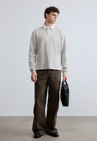 Tânăr îmbrăcat cu un polo bej cu mâneci lungi, pantaloni maro cu croială largă, pantofi negri, ținând o servietă din piele neagră, stând în fața unui fundal simplu, deschis la culoare.