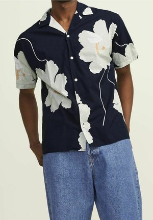 Homme portant une chemise bleu marine à manches courtes avec un grand imprimé floral blanc et un jean en denim bleu clair.