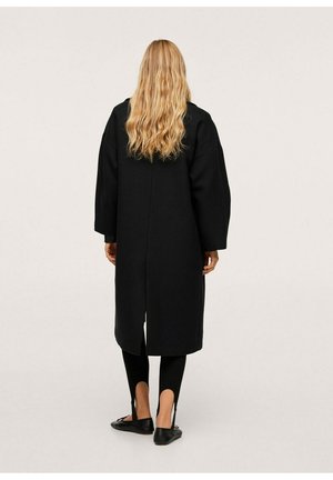 Kobieta z długimi falowanymi blond włosami, ubrana w czarny, oversize płaszcz, czarne legginsy ze strzemionami i czarne płaskie buty, stojąca na jednolitym tle.