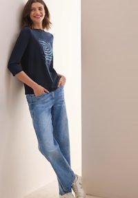 Maglietta blu navy con design grafico, maniche a tre quarti, abbinata a jeans rilassati azzurri e sneakers bianche. Texture lisce visibili.