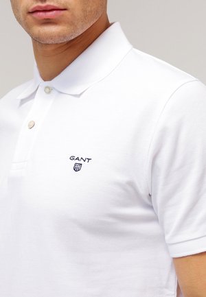 Gros plan d'une personne portant un polo blanc GANT avec une patte de boutonnage à deux boutons et un logo brodé sur la poitrine gauche.