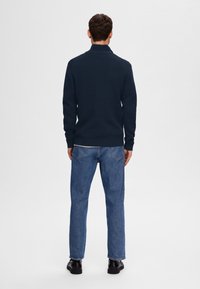 Laivastonsininen neulottu pusero korkealla kauluksella, yhdistetty sinisiin farkkuihin. Asussa on istuvat hihat ja mustat kengät.