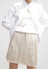 Chemise blanche boutonnée portée sur un débardeur en satin crème et une jupe beige clair à cordon de serrage, longueur genou, avec une texture lisse et des fentes latérales.