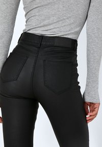 Svarta högmidjade byxor med glansig yta, utrustade med två bakfickor och sömdekor. Matchas med en åtsittande grå långärmad topp.