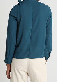 Cyaan lange mouwen blouse met een strik bij de nek, gladde stof en vented achterkant, gecombineerd met crème geribde broek.