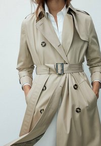 Femme portant un trench-coat beige avec ceinture sur une chemise blanche, les mains dans les poches, mettant en valeur les détails des boutons et le bas du manteau partiellement évasé.