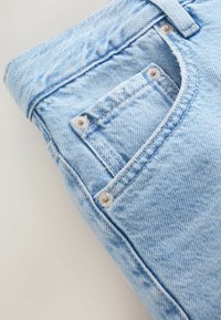 Jean en denim bleu clair plié, montrant la poche avant droite avec petite poche à monnaie et rivets métalliques.