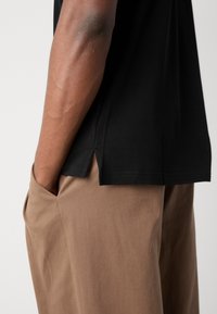 Zwarte katoenen t-shirt met zijopeningen, gecombineerd met bruine stoffen broek. T-shirt heeft een klassieke pasvorm; de broek heeft een relaxed model en zakken.