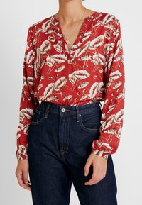 Langarmige, rode blouse met een bloemenpatroon, voorzien van een V-hals en geplooide mouwen, gecombineerd met donkerblauwe high-waisted jeans.